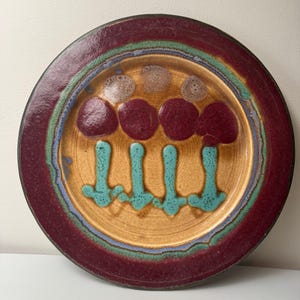 May include: A ceramic plate with a brown, red, and green glaze. The plate features a design of four stylized mushrooms with red caps and turquoise stems.