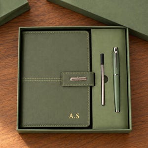 Puede incluir: Un cuaderno verde salvia con cierre magnético y las iniciales A.S. grabadas en oro en la portada, presentado en una caja de regalo a juego junto a una pluma estilográfica verde salvia y un cartucho de tinta en un compartimento.