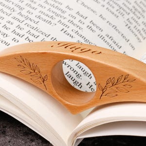May include: A light-colored wooden book page holder with a circular opening. The holder has the name "Hugo" engraved on it, along with leaf designs. It rests on the pages of an open book.