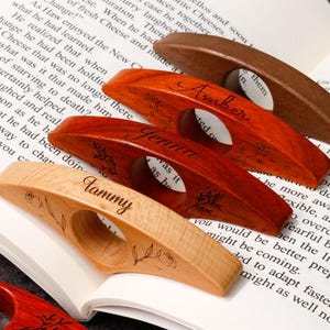May include: Wooden thumb page holders in various wood tones, including light, medium, and dark brown. Each features a circular opening and is engraved with a name and floral design. The names "Tammy" and "Amber" are visible.