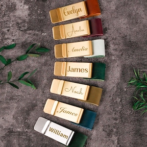 May include: Several personalised name tags with gold-coloured metal plates and leather straps in various colours. Each tag has a different name in elegant script: Evelyn, Amelia, James, Noah, and William. The tags are arranged on a textured grey surface.