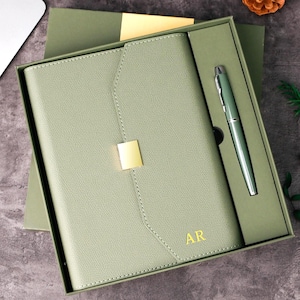 May include: A sage green journal and pen set in a gift box. The journal has a gold clasp and the initials "AR" in gold. The pen is silver. The set is presented on a grey surface with a laptop in the background.