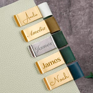 Op de afbeelding: Een lichtgroen leren notitieboek met goud- en zilverkleurige metalen tabs. De tabs zijn gegraveerd met de namen "Amelia", "James" en "Noah". De tabs zijn bevestigd aan de pagina's van het notitieboek, die verschillende kleuren hebben.