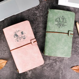 May include: Two personalized journals, one pink and one green, with a pen. The pink journal has the name "Mateo" and the green journal has the name "Michael" engraved on the cover. Both journals are closed with a leather strap.