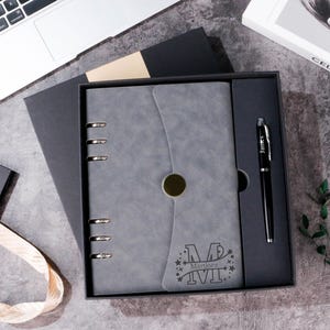 May include: A grey journal and pen set in a black gift box. The journal features a personalised design with the letter "M" and the name "Martinez". A black pen with the name "James" is included. The set is presented on a grey surface.