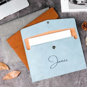 Könnte beinhalten: Drei Laptop-Hüllen aus Leder in Grau, Hellbraun und Hellblau. Die hellblaue Hülle ist geöffnet und zeigt einen weißen Gegenstand im Inneren, mit dem Namen "James" darauf geschrieben. Ein Stift, ein Tannenzapfen und Blätter sind ebenfalls vorhanden.