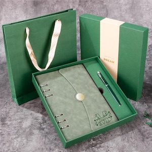Può includere: Un set regalo verde con un diario, una penna e un sacchetto regalo. Il diario è verde oliva con chiusura a bottone e anelli metallici. La penna e la confezione regalo sono anch'esse verdi. Sulla confezione regalo è stampata la parola "DREAM".