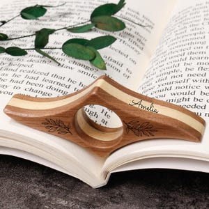 May include: A wooden book page holder with a circular finger hole and the name "Amelia" engraved on it. The holder has a light wood stripe and leaf design. It rests on an open book.