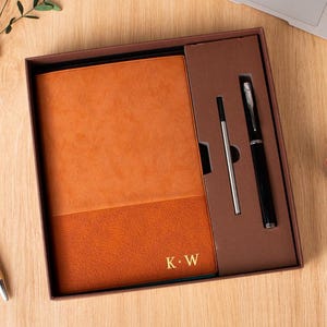 Puede incluir: Una caja de regalo rectangular que contiene un cuaderno naranja con las iniciales K.W grabadas en oro, un bolígrafo negro y un recambio de metal. El cuaderno presenta una cubierta texturizada con un diseño de banda horizontal en dos tonos.