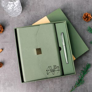 May include: A sage green journal and pen set in a gift box. The journal has a gold square clasp and the name "Violet" with stars. The pen is silver. The box is open, revealing the journal and pen.