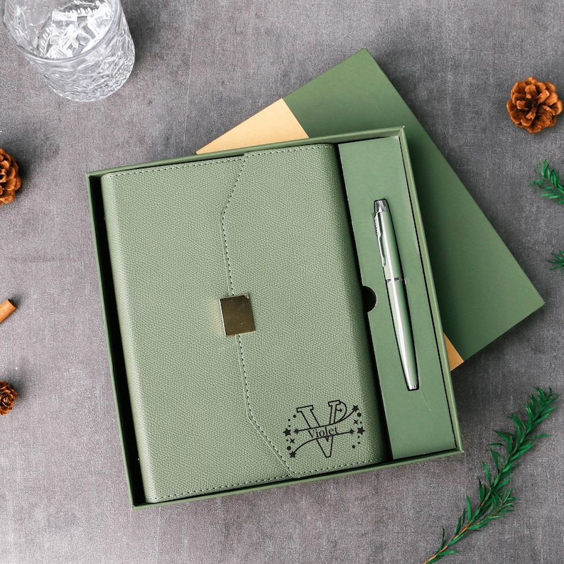 May include: A sage green journal and pen set in a gift box. The journal has a gold square clasp and the name "Violet" with stars. The pen is silver. The box is open, revealing the journal and pen.