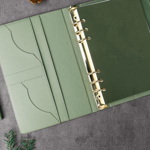 May include: Open view of a sage green leather binder with gold-colored rings and a matching pen. The binder is open, revealing pockets and a dark green interior. The pen is silver and green. The background is a dark gray surface.