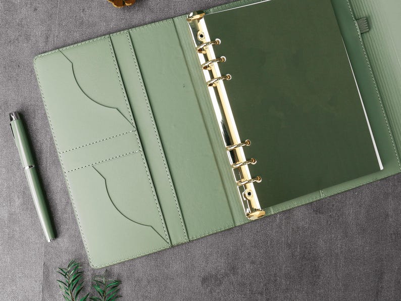 May include: Open view of a sage green leather binder with gold-colored rings and a matching pen. The binder is open, revealing pockets and a dark green interior. The pen is silver and green. The background is a dark gray surface.