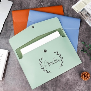 May include: A pale green document folder with the name "Amelia" in script, encircled by a laurel wreath design. Other folders are orange and blue. A laptop and a book are also visible.