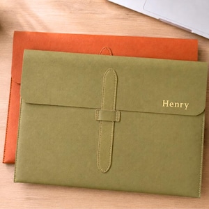 May include: An olive green document folder with a vertical strap closure and the name Henry embossed in gold on the front flap, layered over a larger orange document folder. Both folders feature a smooth, matte finish and stitched edges.