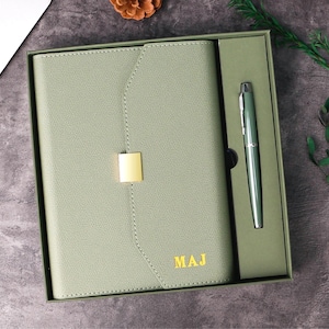 Puede incluir: Un juego de diario y bolígrafo verde salvia en una caja de regalo. El diario tiene un cierre dorado y las iniciales "MAJ" en dorado. El bolígrafo es plateado y verde. El juego se presenta sobre una superficie gris oscuro con un portátil y una piña en el fondo.