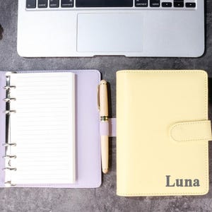 Pode incluir: Um planejador amarelo claro com o nome "Luna" impresso na frente, ao lado de um planejador lilás aberto com papel pautado. Uma caneta dourada e lilás repousa entre os dois planejadores. Um laptop e um pinha estão no fundo.