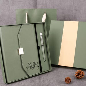May include: A sage green gift set featuring a notebook, pen, and gift box. The notebook has a flap closure with a silver clasp and the word "Violet" printed on it. A matching gift bag and two pinecones complete the presentation.