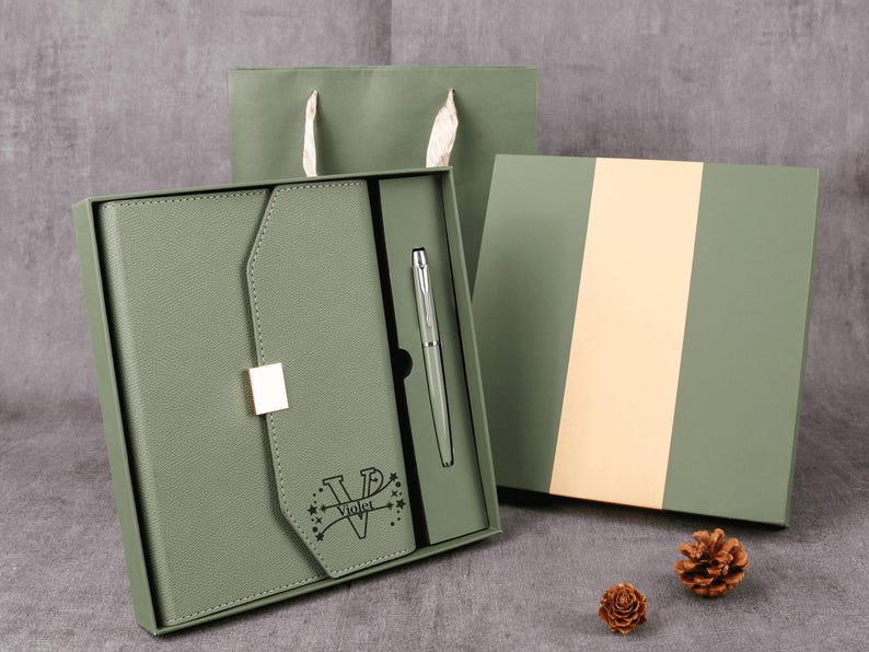 May include: A sage green gift set featuring a notebook, pen, and gift box. The notebook has a flap closure with a silver clasp and the word "Violet" printed on it. A matching gift bag and two pinecones complete the presentation.