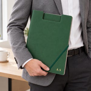 May include: A dark green document folder with a rectangular cutout handle at the top, secured by a matching elastic band. The bottom right corner features the gold embossed initials A.S. The folder has a smooth, solid-coloured exterior finish.