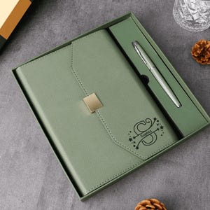 May include: Sage green journal and pen set in a gift box. The journal has a gold-colored square clasp and the name "Sophia" is printed on the cover. A silver pen is included in the box.