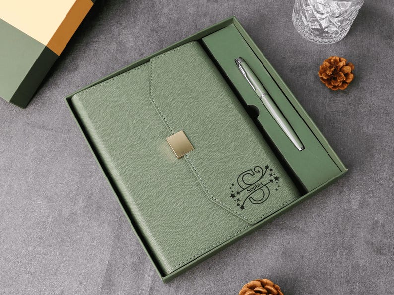 May include: Sage green journal and pen set in a gift box. The journal has a gold-colored square clasp and the name "Sophia" is printed on the cover. A silver pen is included in the box.