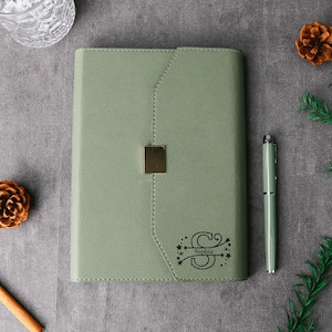 May include: Sage green journal with a gold-colored clasp and matching pen. The journal is personalized with the name "Sophia" and a decorative initial "S". The journal is surrounded by decorative pinecones and greenery.