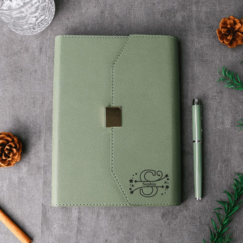 May include: Sage green journal with a gold-colored clasp and matching pen. The journal is personalized with the name "Sophia" and a decorative initial "S". The journal is surrounded by decorative pinecones and greenery.