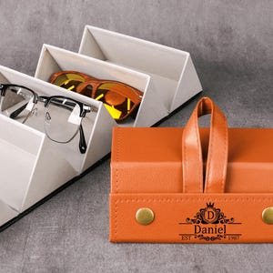 Puede incluir: Un estuche de gafas de cuero naranja con asa y cierres de botón dorados. El estuche está personalizado con el nombre "Daniel" y el año "1987". Gafas blancas y negras y gafas de sol naranjas se exhiben en un soporte blanco con múltiples compartimentos.