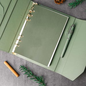 May include: An open, sage green leather binder with gold-colored rings and a matching pen. The binder is lying open on a gray surface, with a cinnamon stick, pine cone, and sprig of greenery nearby.