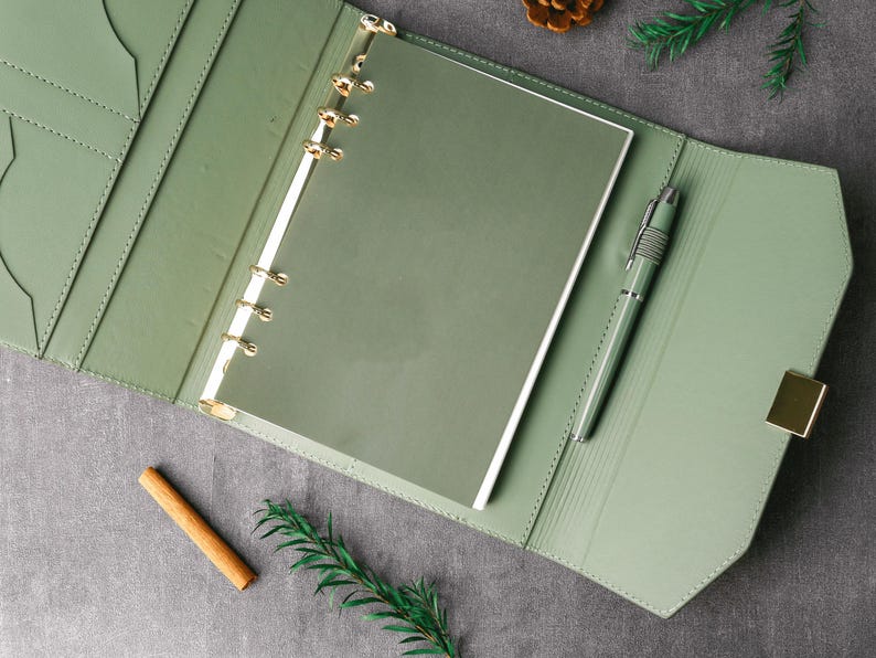 May include: An open, sage green leather binder with gold-colored rings and a matching pen. The binder is lying open on a gray surface, with a cinnamon stick, pine cone, and sprig of greenery nearby.