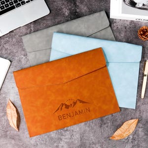 May include: Three leather document holders in tan, grey, and light blue are displayed on a grey surface. The tan holder has a mountain graphic and the name "BENJAMIN" printed on it. A laptop, notebook, pen, and decorative items are also present.