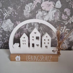 Könnte beinhalten: Weißer Holzausschnitt von drei Häusern mit Herzen und einem Stern, unter einem Halbkreisbogen. Die Häuser stehen auf einem Holzsockel mit dem Text "LIEBLINGSPLATZ" in Weiß.