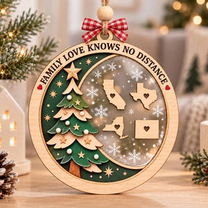 Op de afbeelding: Een ronde houten kerstornament met een gelaagde groene boom met sterren en witte bollen, een doorzichtig gedeelte met vier Amerikaanse staatskaarten met hartvormige uitsparingen en de tekst FAMILY LOVE KNOWS NO DISTANCE.