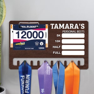 May include: A wooden medal display board with the text "TAMARA'S PERSONAL BESTS" and slots for 5K, 10K, Half, and Full marathon times. It also features a race bib with the number 12000 and several colorful marathon ribbons.