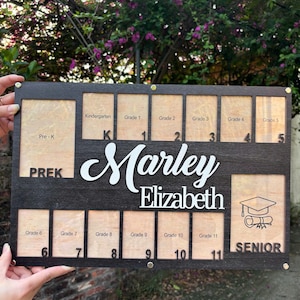 May include: A personalized wooden school years photo display board. The black board has spaces for photos from Pre-K to Grade 11, with the name "Marley Elizabeth" in white. A graduation cap and scroll are displayed in the Senior section.