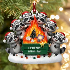 May include: A festive Christmas tree ornament depicting six raccoons in Santa hats gathered around a dumpster fire. The dumpster has the text "DUMPSTER FIRE RESPONSE TEAM". Includes a small Christmas tree and presents.