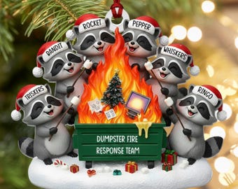 May include: A Christmas ornament featuring five raccoons wearing Santa hats labeled Bandit, Rocket, Pepper, Whiskers, and Ringo. They stand around a green dumpster fire labeled DUMPSTER FIRE RESPONSE TEAM, holding sticks with marshmallows.