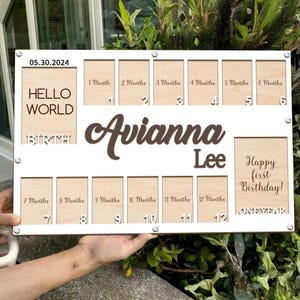 Könnte beinhalten: Eine weiße Meilensteintafel aus Holz mit Fotorahmen. Die Tafel enthält den Namen "Avianna Lee", den Text "Hello World" und "Happy first Birthday!" sowie das Datum 05.30.2024.
