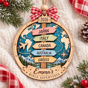 May include: A round wooden Christmas ornament featuring a world map background, a vertical post with five directional signs labeled Japan, Italy, Canada, Australia, and Greece, a 2026 tag, a red gingham bow, and the text Emma's WORLD ADVENTURES.