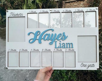 Puede incluir: Marco de fotos de madera blanco con 13 ranuras para fotos. El marco está diseñado para el primer año de un bebé, con etiquetas para cada mes y "Un año". Los nombres "Hayes Liam" están escritos en letras cursivas azules.