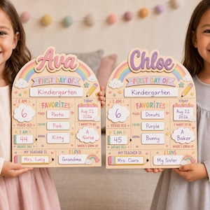 May include: Two arched wooden signs for the first day of kindergarten, personalized with the names Ava and Chloe. Each sign features a rainbow, clouds, and sections for age, height, favorites, date, teacher, and future career goals in colorful text.