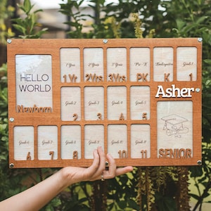 May include: Wooden school years photo display board with the name "Asher". The board has spaces for photos from newborn to senior year, with text like "Hello World" and "Newborn".
