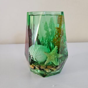 May include: A green glass vase with a geometric shape filled with sea life, including a starfish, a seashell, and small pebbles.