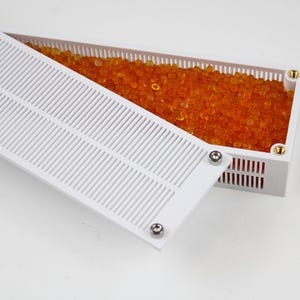 Könnte beinhalten: Ein weißer Plastikbehälter mit einem abnehmbaren Deckel, gefüllt mit orangefarbenen Silicagel-Kügelchen. Der Deckel hat ein Gittermuster und ist mit kleinen Schrauben befestigt.