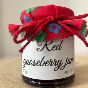 May include: A glass jar of red gooseberry jam with a white label featuring the text Red gooseberry jam, 227g, and BBE 09/27. The lid is covered with a red fabric featuring a green and blue floral pattern, secured with a red string tied in a bow.