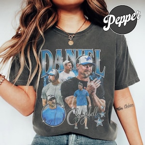 Peut inclure: Un t-shirt gris anthracite à manches courtes avec une impression graphique de Daniel Campbell. Le motif comprend plusieurs images de l'entraîneur, le nom DANIEL en grandes lettres bleues et plusieurs étoiles. Le t-shirt a un col rond.