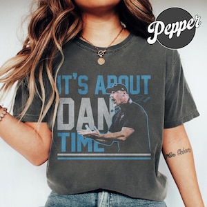 Peut inclure: Un t-shirt gris anthracite à manches courtes avec un graphique représentant un homme portant une casquette de baseball et le texte IT'S ABOUT DAN TIME en bleu clair et blanc. Le t-shirt a un col rond et une finition en tissu délavé.