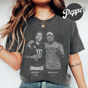 May include: A charcoal grey short-sleeve t-shirt featuring a graphic print of Jared Goff and Dan Campbell. The design includes the text "Jared Goff" and "Dan Campbell" below the figures, with "Detroit" printed on the shirt worn by Campbell.