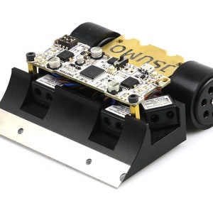 Op de afbeelding: Een kleine, zwart-zilveren robot met een printplaat en het woord "SUMO" op een gouden plaat. De robot heeft twee zwarte cilindrische wielen en een zilveren mes aan de voorkant.